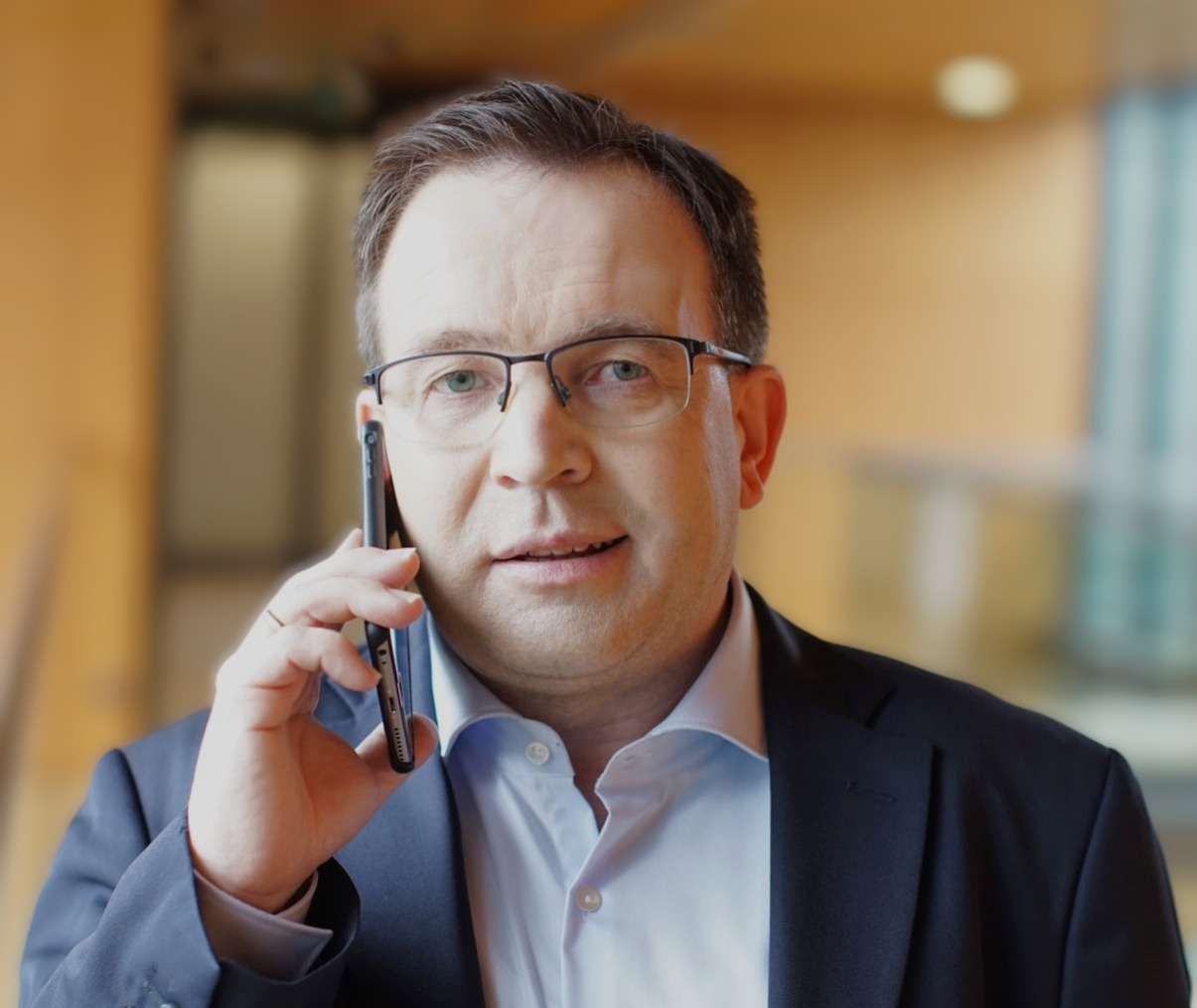 Mężczyzna w okularach i garniturze rozmawia przez telefon, uśmiechając się lekko. Jasne tło, biznesowy charakter zdjęcia.