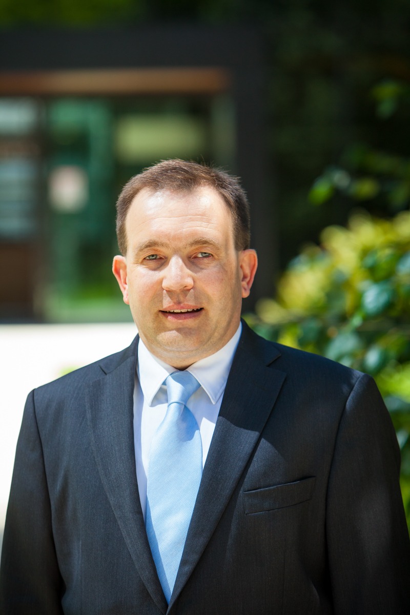 Portret mężczyzny w garniturze z jasnoniebieskim krawatem, uśmiechającego się do kamery na tle zieleni i budynku. Dobre oświetlenie, profesjonalny wygląd.