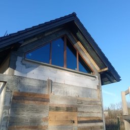 DUET SPÓŁKA Z OGRANICZONĄ ODPOWIEDZIALNOŚCIĄ - Nietypowe okno trójkątne z brązową ramą w surowym stanie budynku, widok z dołu na tle błękitnego nieba.