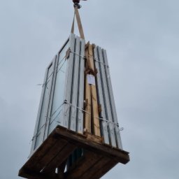 DUET SPÓŁKA Z OGRANICZONĄ ODPOWIEDZIALNOŚCIĄ - Paczka okien w białych ramach zabezpieczona na drewnianej palecie, podnoszona przez żuraw na tle pochmurnego nieba.