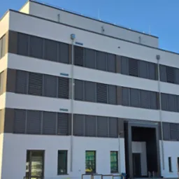 Nowoczesny budynek biurowy z szarymi roletami zewnętrznymi na wszystkich oknach, widoczne rynny spustowe, błękitne niebo w tle.