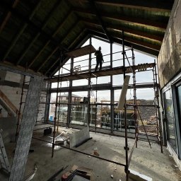 DUET SPÓŁKA Z OGRANICZONĄ ODPOWIEDZIALNOŚCIĄ - Montaż dużych okien w surowym stanie budynku, pracownik na rusztowaniu, widok z wnętrza, konstrukcja dachu z drewnianymi belkami.