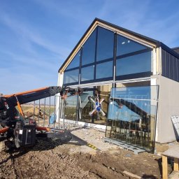 DUET SPÓŁKA Z OGRANICZONĄ ODPOWIEDZIALNOŚCIĄ - Montaż okien aluminiowych w nowoczesnym domu z użyciem dźwigu przyssawkowego, widoczne szyby i rusztowania.