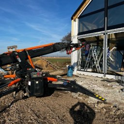 DUET SPÓŁKA Z OGRANICZONĄ ODPOWIEDZIALNOŚCIĄ - Czarny dźwig pająk z pomarańczowymi elementami podnosi szybę na tle nowoczesnego domu z dużymi oknami, widoczne drabiny i pracownik wewnątrz budynku.