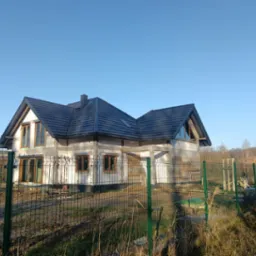 Dom w trakcie budowy z zamontowanymi oknami z drewnianą ramą, widok zza ogrodzenia z siatki.