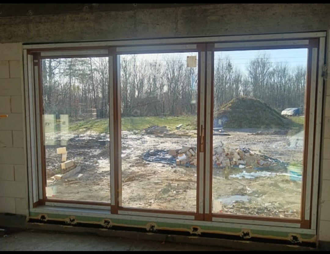 Zamontowane trzyskrzydłowe drzwi przesuwne z brązowymi ramami w surowym stanie budynku, widok na niezagospodarowany teren z hałdą ziemi.