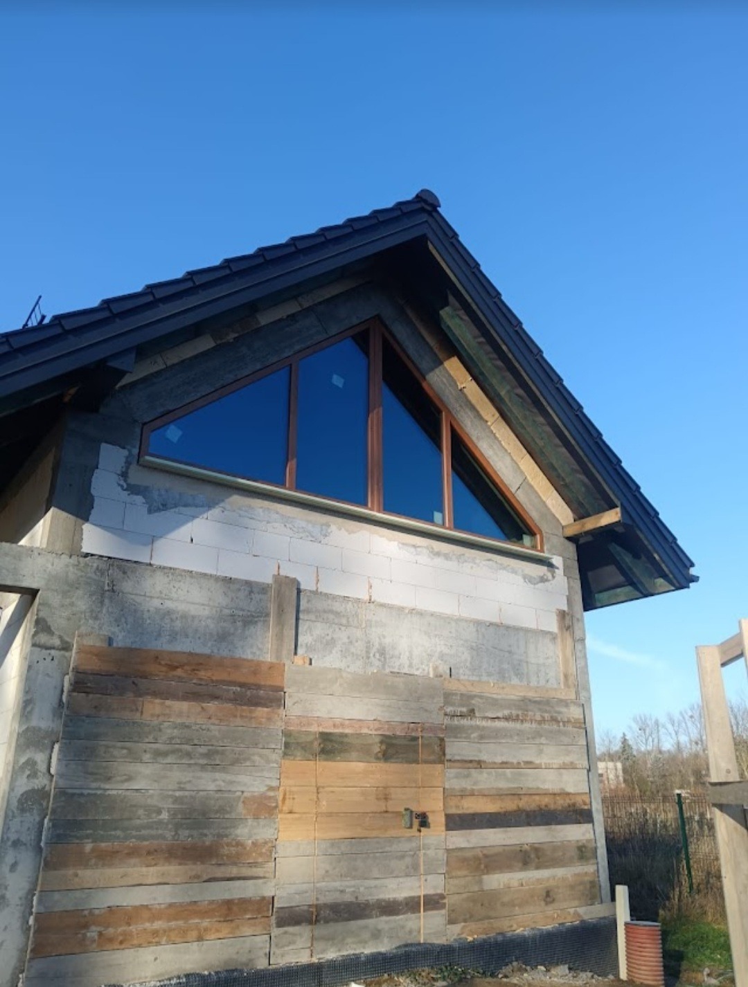 Nietypowe okno trójkątne z brązową ramą w surowym stanie budynku, widok z dołu na tle błękitnego nieba.