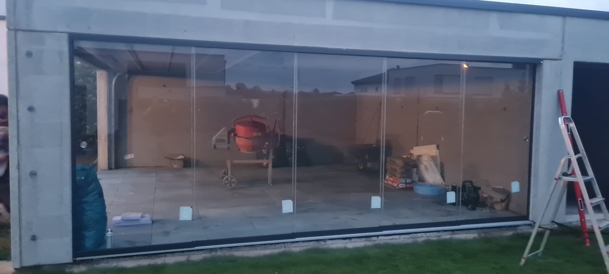 Szklane drzwi przesuwne w betonowej konstrukcji garażu, widoczny mikser do betonu i drabina, w tle dom.