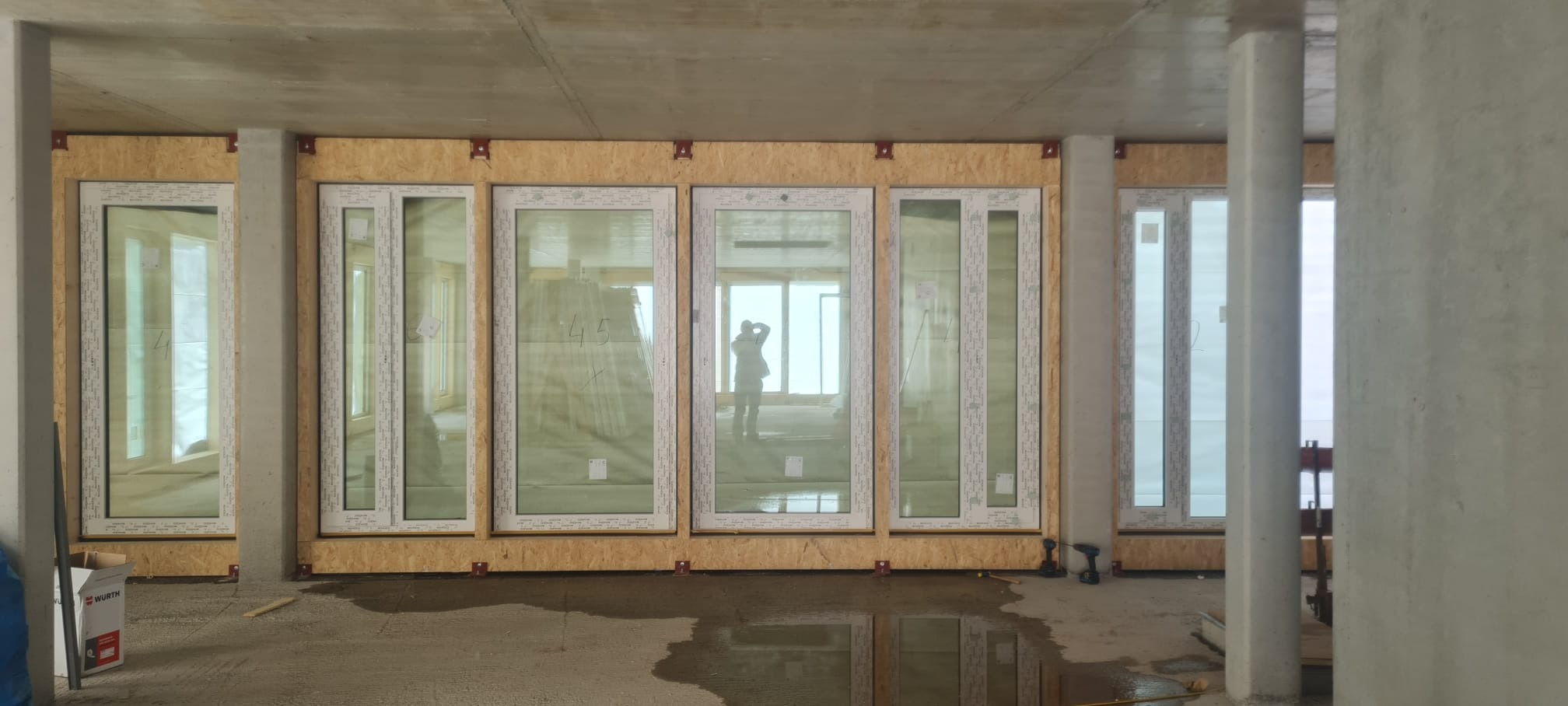 Montaż okien w surowym stanie: białe ramy okienne wstawione w drewniane płyty, betonowe ściany i podłoga z kałużami, sylwetka fotografa w odbiciu szyby.