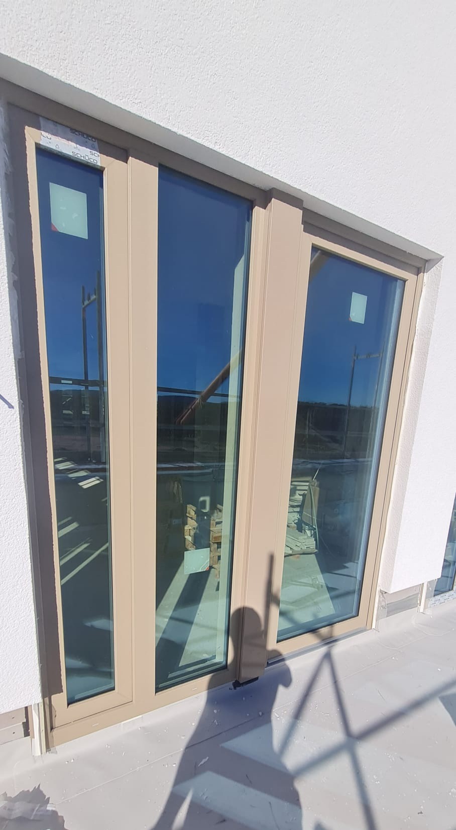 Zamontowane, beżowe okno z trzema szybami w nowym budynku, widoczne elementy konstrukcyjne i palety z materiałami wewnątrz.