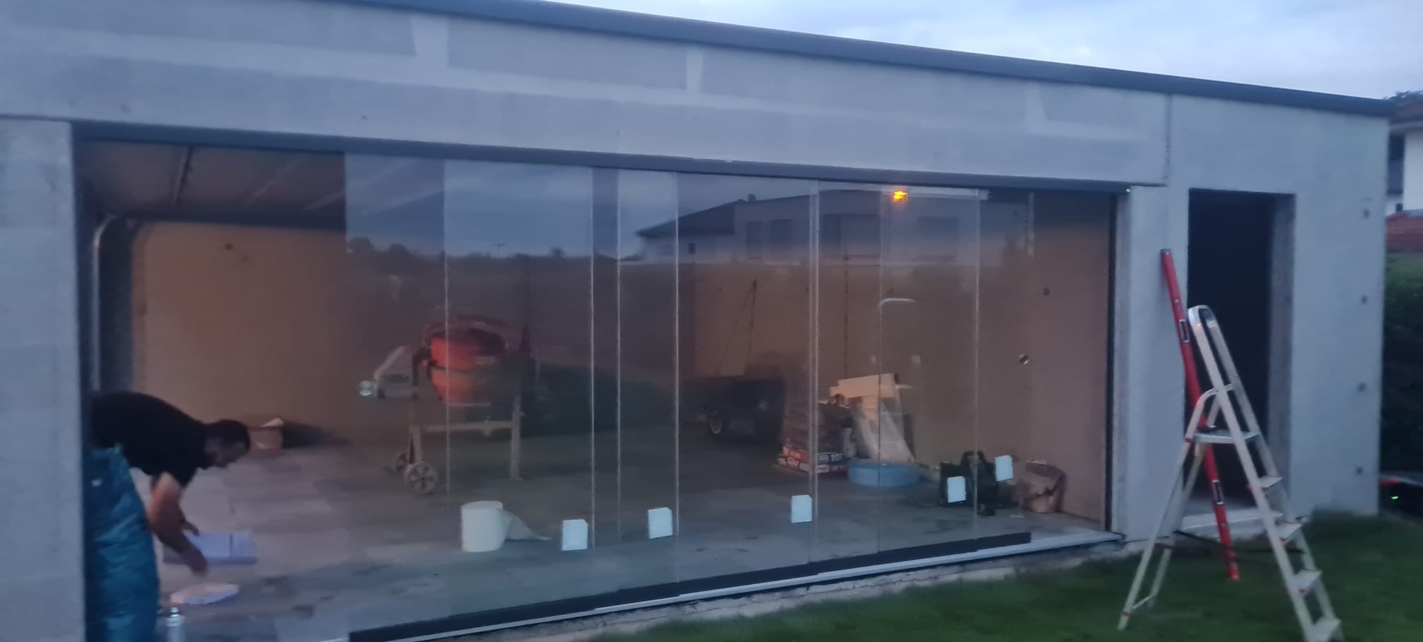 Montaż drzwi przesuwnych szklanych w garażu; widoczna drabina, poziomica i pracownik przygotowujący się do pracy.