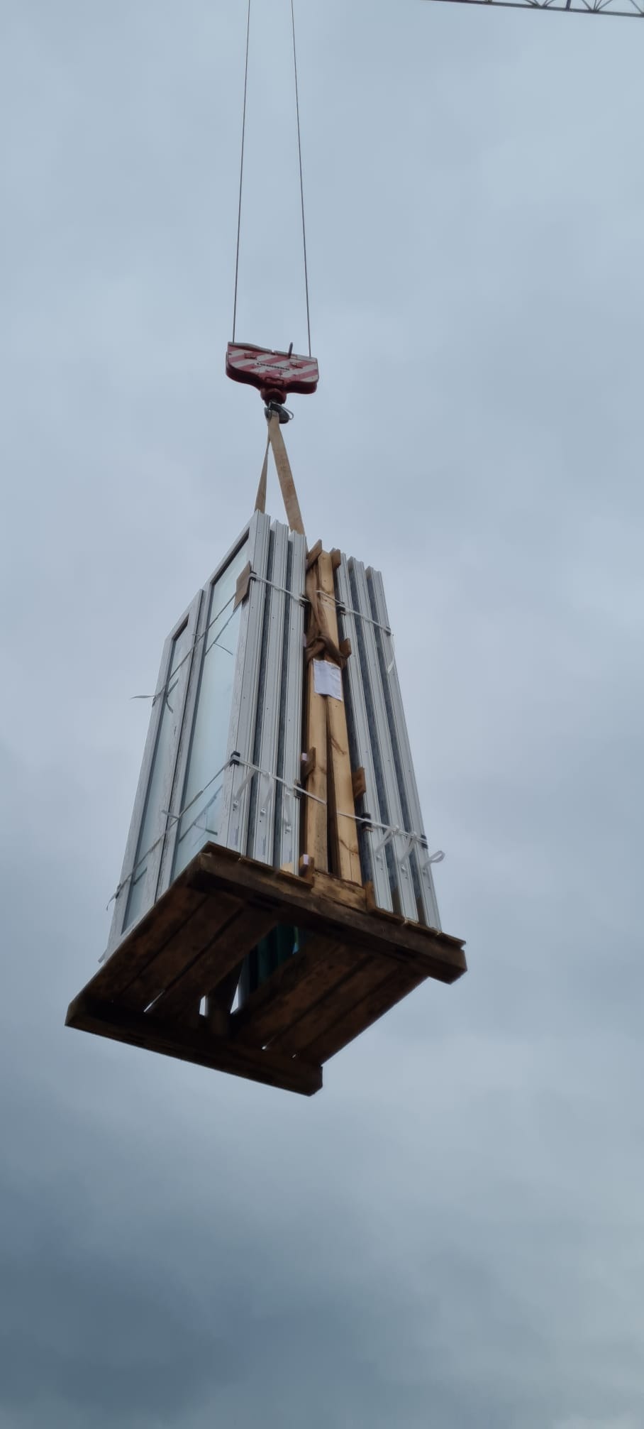 Paczka okien w białych ramach zabezpieczona na drewnianej palecie, podnoszona przez żuraw na tle pochmurnego nieba.