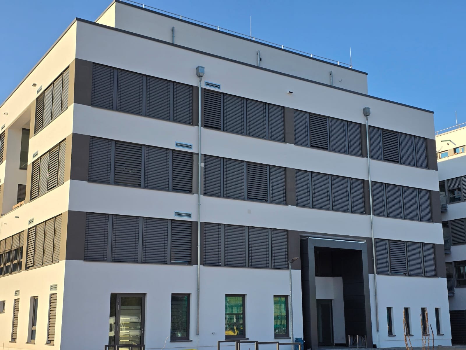 Nowoczesny budynek biurowy z szarymi roletami zewnętrznymi na wszystkich oknach, widoczne rynny spustowe, błękitne niebo w tle.