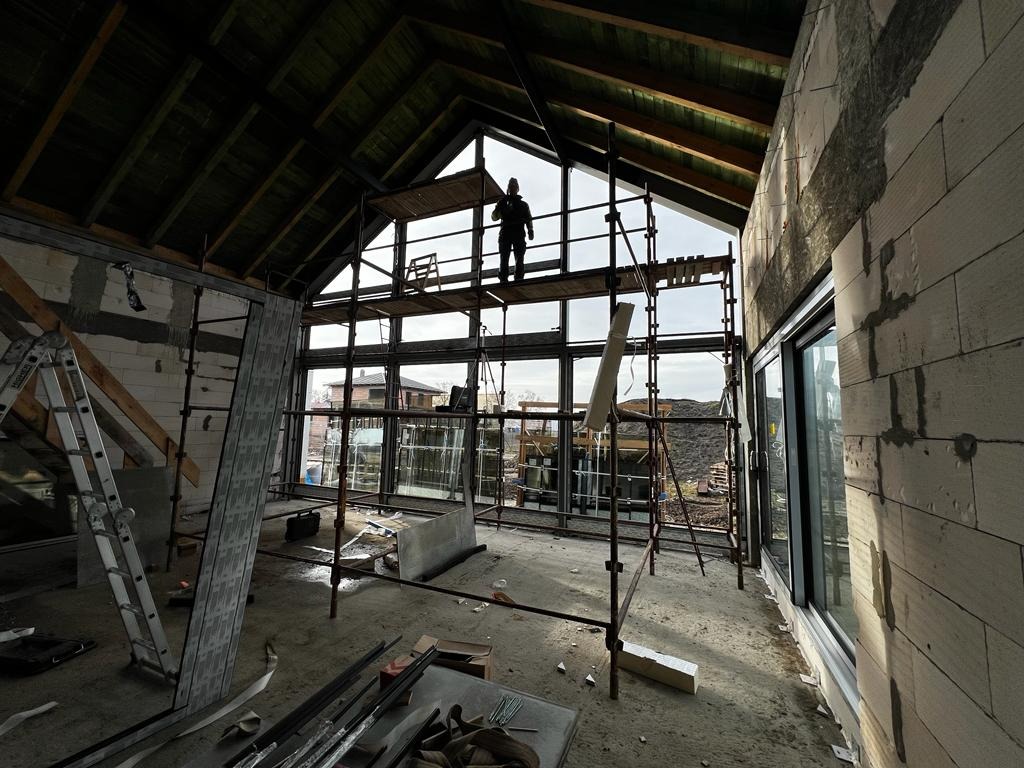 Wnętrze surowego domu w trakcie budowy z widocznymi rusztowaniami, niezamontowanymi oknami i pracownikiem na rusztowaniu, z surowym drewnianym dachem i ścianami z betonu komórkowego.