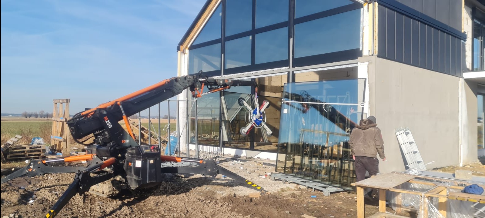 Montaż dużych, panoramicznych okien w nowym domu za pomocą specjalistycznego dźwigu próżniowego; widoczny pracownik w trakcie pracy, przygotowane tafle szkła i rusztowania.