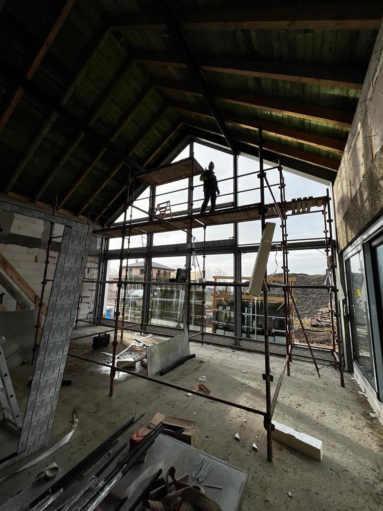 Montaż dużych okien w surowym stanie budynku, pracownik na rusztowaniu, widok z wnętrza, konstrukcja dachu z drewnianymi belkami.