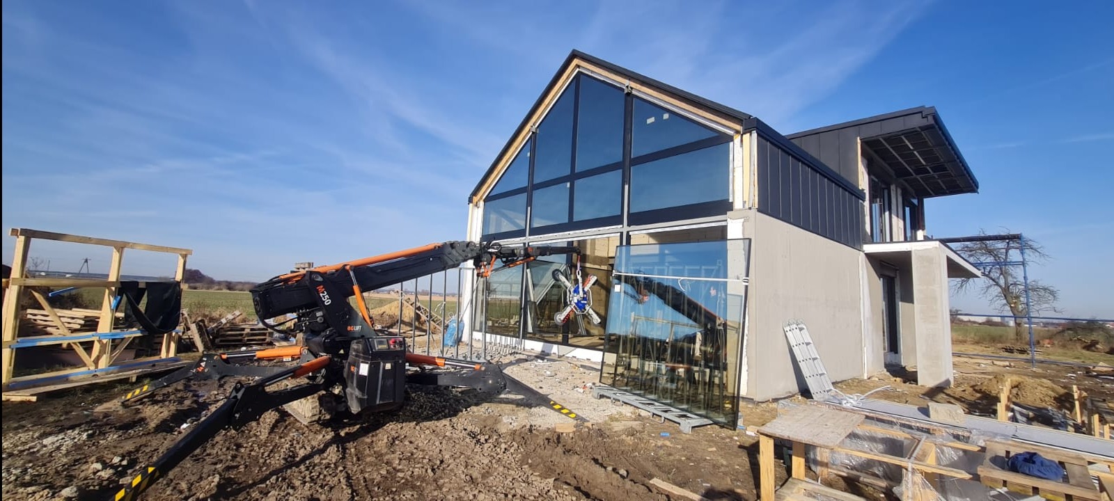 Montaż okien aluminiowych w nowoczesnym domu z użyciem dźwigu przyssawkowego, widoczne szyby i rusztowania.