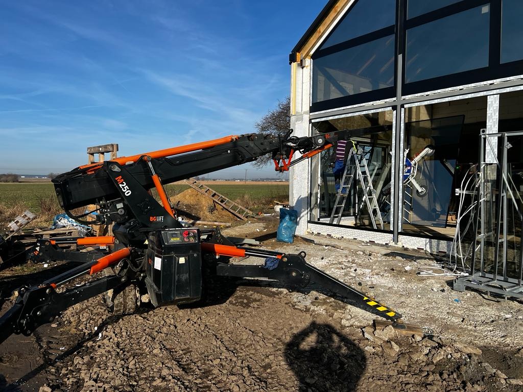 Czarny dźwig pająk z pomarańczowymi elementami podnosi szybę na tle nowoczesnego domu z dużymi oknami, widoczne drabiny i pracownik wewnątrz budynku.