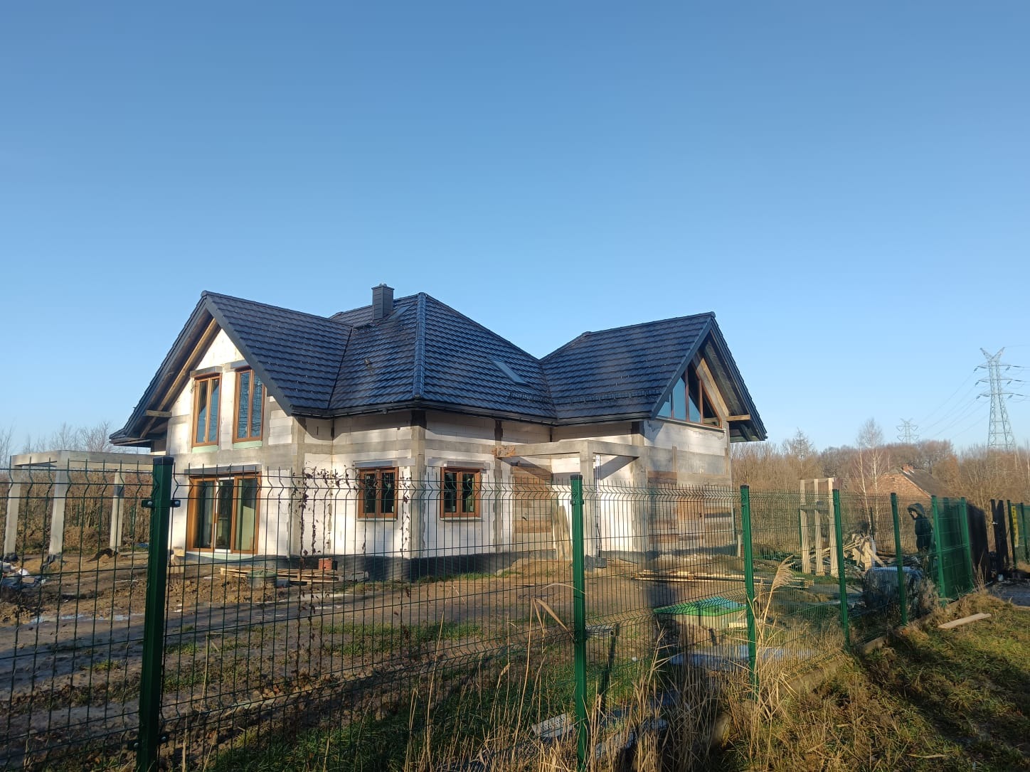 Dom w trakcie budowy z zamontowanymi oknami z drewnianą ramą, widok zza ogrodzenia z siatki.