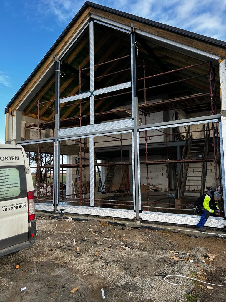 Montaż dużych okien w surowym stanie budynku, widoczne rusztowania wewnątrz, pracownik w odblaskowej kurtce przy pracy, na zewnątrz zaparkowany bus z logo firmy.