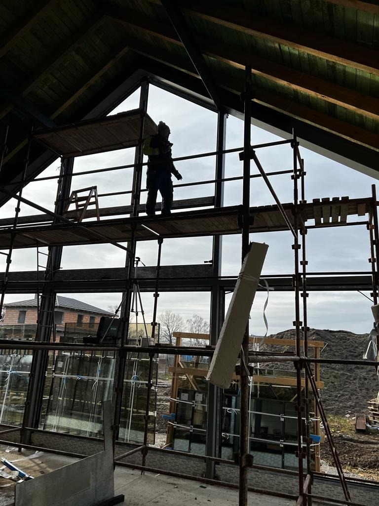 Pracownik na rusztowaniu montuje duże okno w budynku z drewnianą konstrukcją dachu, widok na zewnątrz przez niezamontowane jeszcze szyby.