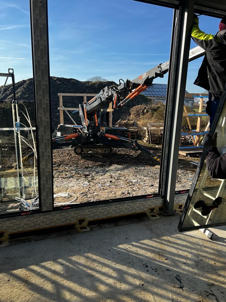 Montaż okna z użyciem mini-żurawia na gąsienicach, widoczny pracownik na drabinie w trakcie wstawiania szyby, w tle hałda ziemi i konstrukcja szkieletowa budynku.