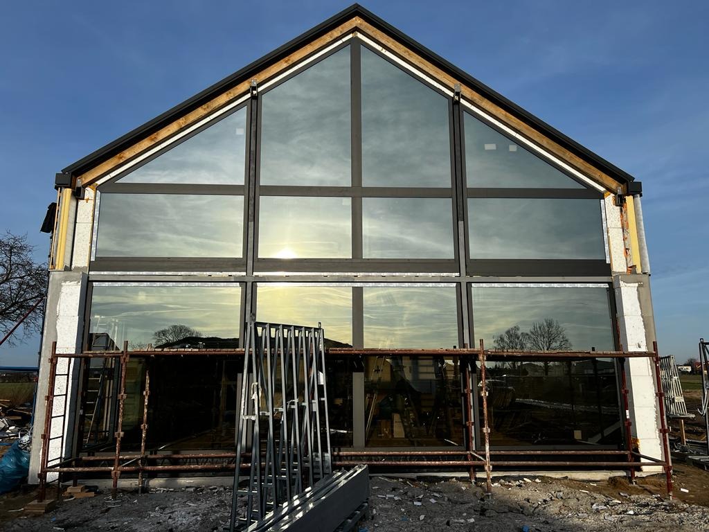 Nowoczesny dom w trakcie budowy z dużymi, panoramicznymi oknami z czarnymi ramami, widoczne rusztowania i materiały budowlane.