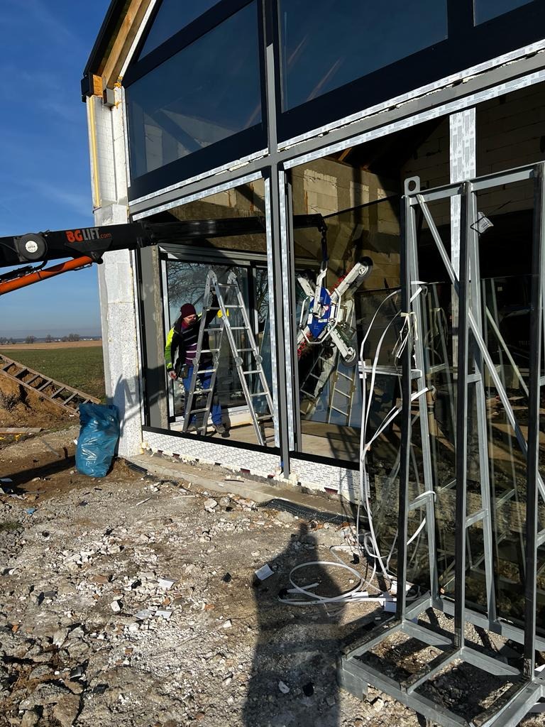Montaż dużych okien aluminiowych w surowym stanie budynku, widoczny dźwig z przyssawkami do szkła, drabina i pracownik w odblaskowej kamizelce.