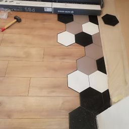 ATOM ADAM SROKA - Fragment podłogi z łączonym wzorem: drewniane panele i heksagonalne płytki w odcieniach bieli, szarości i czerni. Widoczne opakowania listew przypodłogowych Kronopol Platinum P85 oraz gumowy młotek.