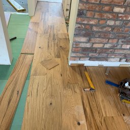 ATOM ADAM SROKA - Układanie drewnianej podłogi w korytarzu z ceglaną ścianą, widoczne narzędzia i fragmenty paneli na zielonym podkładzie.