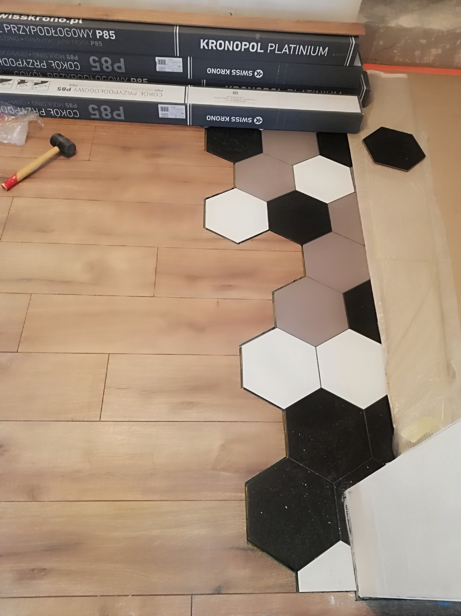 Fragment podłogi z łączonym wzorem: drewniane panele i heksagonalne płytki w odcieniach bieli, szarości i czerni. Widoczne opakowania listew przypodłogowych Kronopol Platinum P85 oraz gumowy młotek.