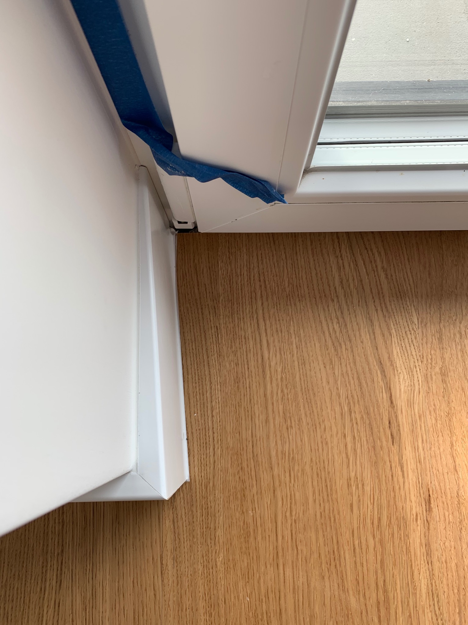 Fragment białej ościeżnicy z niebieską taśmą malarską zabezpieczającą, przylegająca do drewnianej podłogi.