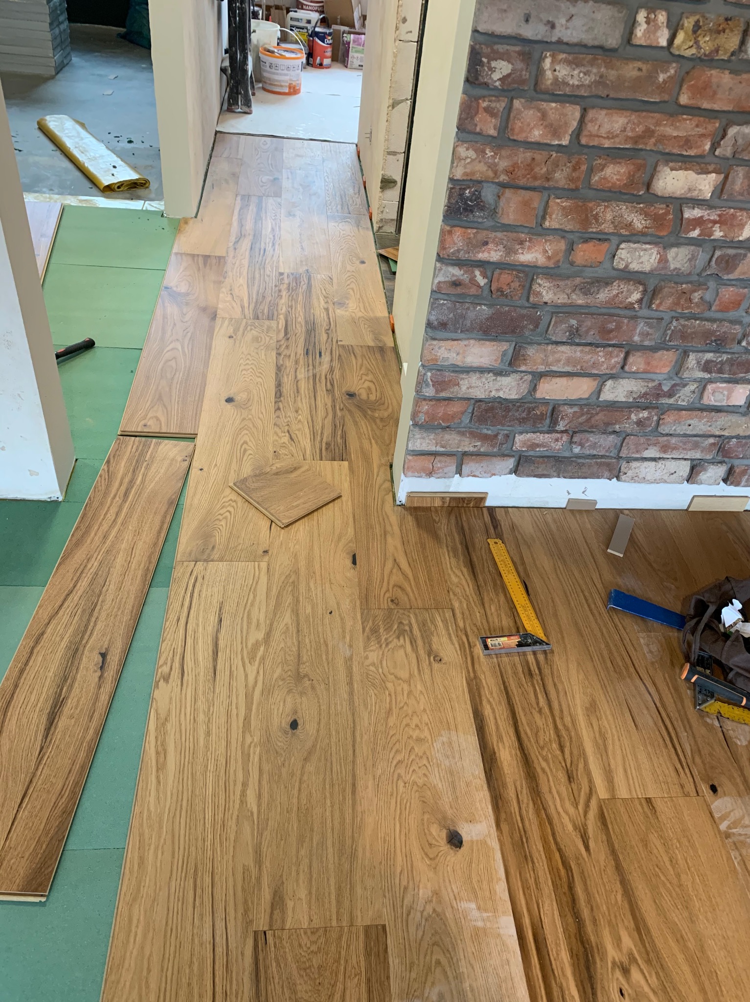 Układanie drewnianej podłogi w korytarzu z ceglaną ścianą, widoczne narzędzia i fragmenty paneli na zielonym podkładzie.
