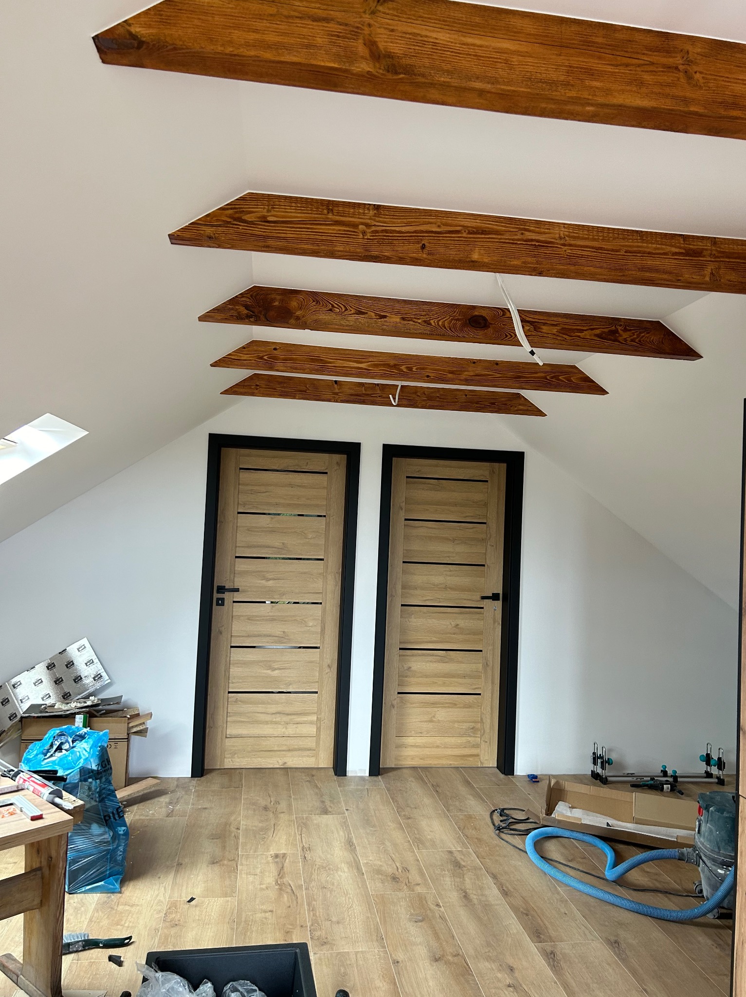 Poddasze po remoncie: dwie drewniane drzwi z czarnymi ościeżnicami, drewniane belki stropowe, panele podłogowe i widoczne narzędzia pracy.