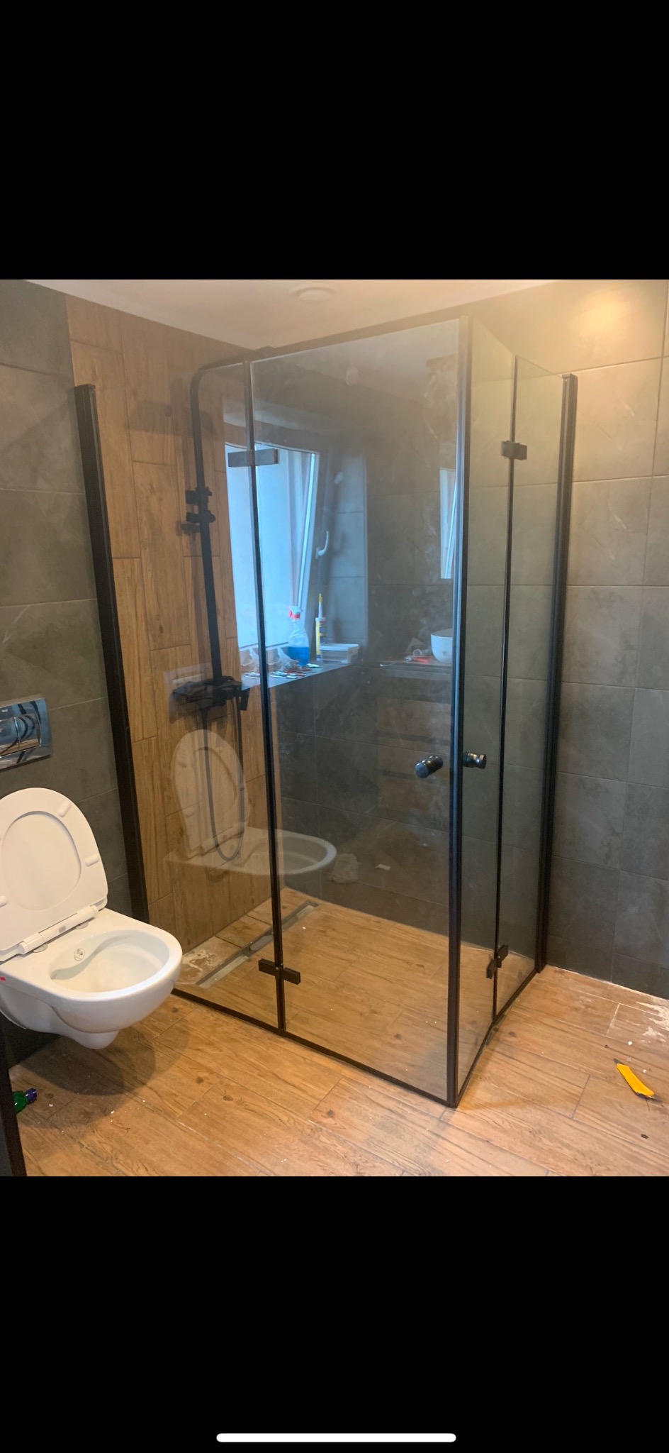 Nowoczesna kabina prysznicowa z czarnymi profilami i szklanymi ściankami, zamontowana w łazience z drewnopodobną podłogą i szarymi płytkami ściennymi. Widoczna wisząca muszla klozetowa z otwartą...