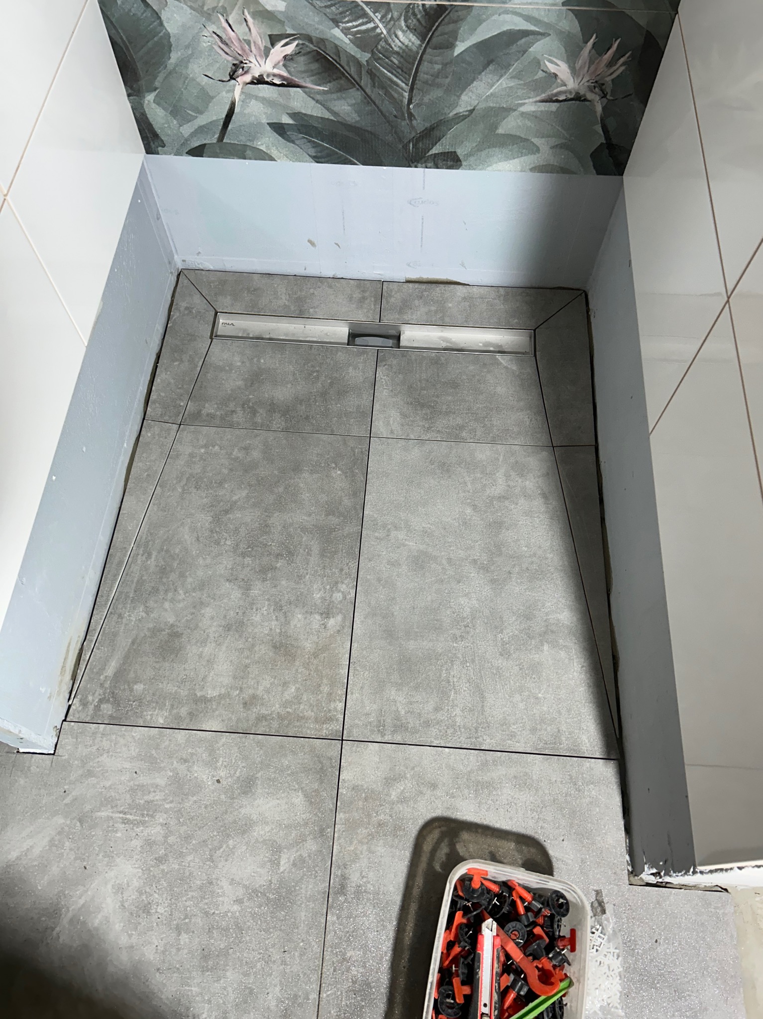 Szare płytki podłogowe ułożone w kabinie prysznicowej typu walk-in, widoczny odpływ liniowy i narzędzia do układania płytek.