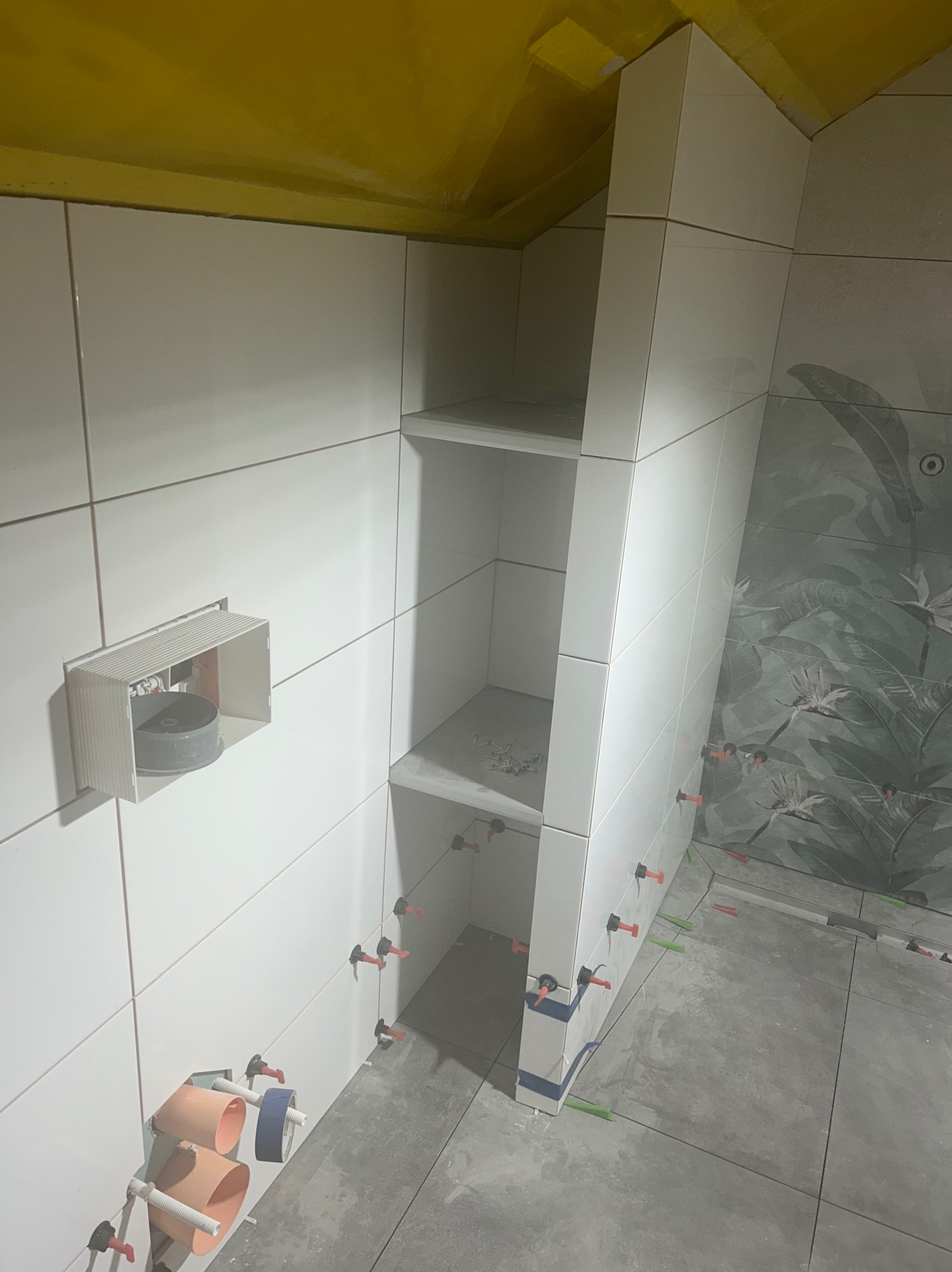 Łazienka w trakcie wykańczania: białe płytki na ścianach z wnęką z półkami, płytki podłogowe w odcieniach szarości, widoczne narzędzia i elementy montażowe, ściana z motywem roślinnym w tle.