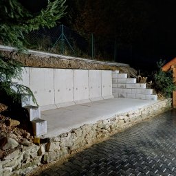 TAKIN USŁUGI BUDOWLANE DAWID JASKÓŁKA - Mur oporowy z betonowych elementów z niskimi schodami, podtrzymujący skarpę, wykończony kamienną podmurówką, widok na mokrą kostkę brukową i ogrodzenie z siatki w tle, nocne oświetlenie.