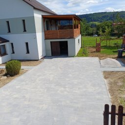 TAKIN USŁUGI BUDOWLANE DAWID JASKÓŁKA - Nowo ułożona, jasnoszara kostka brukowa na podjeździe przed nowoczesnym, dwupiętrowym domem z garażem i drewnianym balkonem, widok z góry na świeżo wykonaną nawierzchnię.