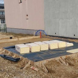 TAKIN USŁUGI BUDOWLANE DAWID JASKÓŁKA - Przygotowanie do izolacji fundamentów: ułożona folia hydroizolacyjna w drewnianym szalunku, obok stosy płyt izolacyjnych Swissopor, w tle budynki mieszkalne i rury instalacyjne.