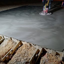 TAKIN USŁUGI BUDOWLANE DAWID JASKÓŁKA - Świeżo wylany beton wyrównywany maszyną zacieraczką, widoczne drewniane szalunki i przygotowane podłoże z kamieni i ziemi, nocne oświetlenie budowy