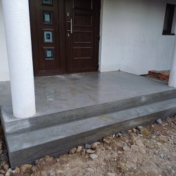 TAKIN USŁUGI BUDOWLANE DAWID JASKÓŁKA - Nowy, betonowy podest wejściowy z dwoma stopniami, wykończony przy białych kolumnach i brązowych drzwiach wejściowych. Otoczenie w trakcie zagospodarowania.