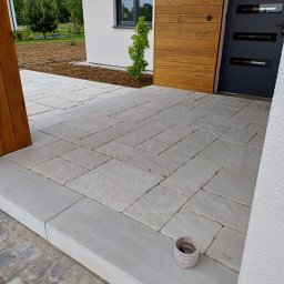 TAKIN USŁUGI BUDOWLANE DAWID JASKÓŁKA - Jasnoszara kostka brukowa ułożona na podjeździe domu z drewnianą elewacją. Widoczny fragment rabaty i doniczka. Ujęcie z bliska, podkreślające wzór i fakturę kostki.