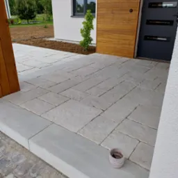 Jasnoszara kostka brukowa ułożona na podjeździe domu z drewnianą elewacją. Widoczny fragment rabaty i doniczka. Ujęcie z bliska, podkreślające wzór i fakturę kostki.