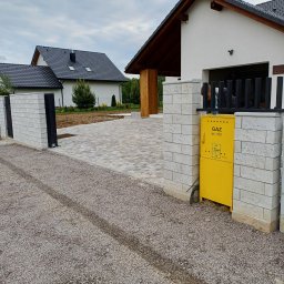 TAKIN USŁUGI BUDOWLANE DAWID JASKÓŁKA - Nowoczesny podjazd z kostki brukowej prowadzący do domu z garażem, widoczna brama wjazdowa z elementami ogrodzenia i skrzynka gazowa.