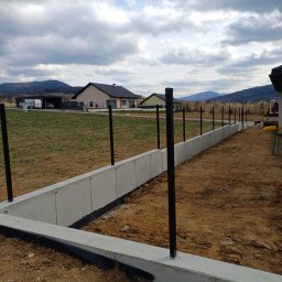 TAKIN USŁUGI BUDOWLANE DAWID JASKÓŁKA - Betonowa podmurówka ogrodzenia z czarnymi słupkami wkopanymi w ziemię. W tle domy i góry pod pochmurnym niebem.