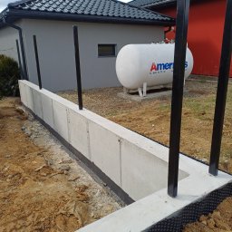 TAKIN USŁUGI BUDOWLANE DAWID JASKÓŁKA - Betonowa podmurówka ogrodzenia z czarnymi słupkami na tle domu z zbiornikiem gazu. Widoczne zabezpieczenie hydroizolacyjne fundamentu.