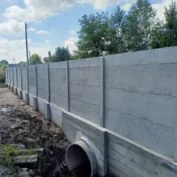 Długi, betonowy płot panelowy z rurą drenażową w ziemi, widok z boku. Tło: niebo z chmurami i drzewa. Elementy są szare, ziemia brązowa, a niebo błękitne.