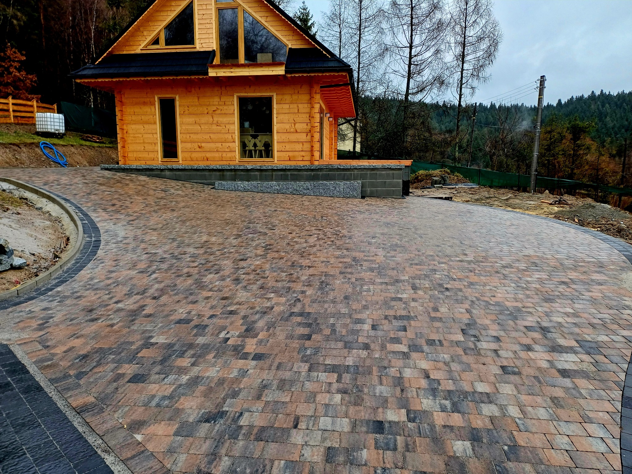 Szeroki podjazd z kostki brukowej w odcieniach brązu i szarości prowadzący do drewnianego domu z czarnym dachem, na tle zalesionego wzgórza i pochmurnego nieba.