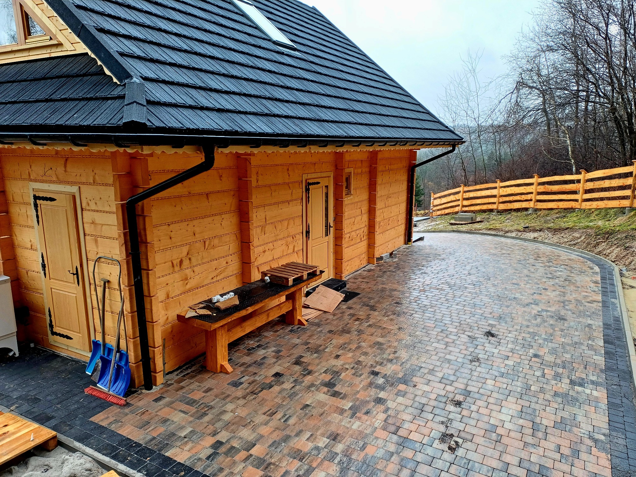 Nowo ułożona, wielobarwna kostka brukowa prowadząca do drewnianego domku z czarnym dachem; w tle widoczne drewniane ogrodzenie i drzewa.